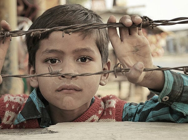 A young boy gazes through a barbed wire fence, his curious eyes filled with longing and innocence.
